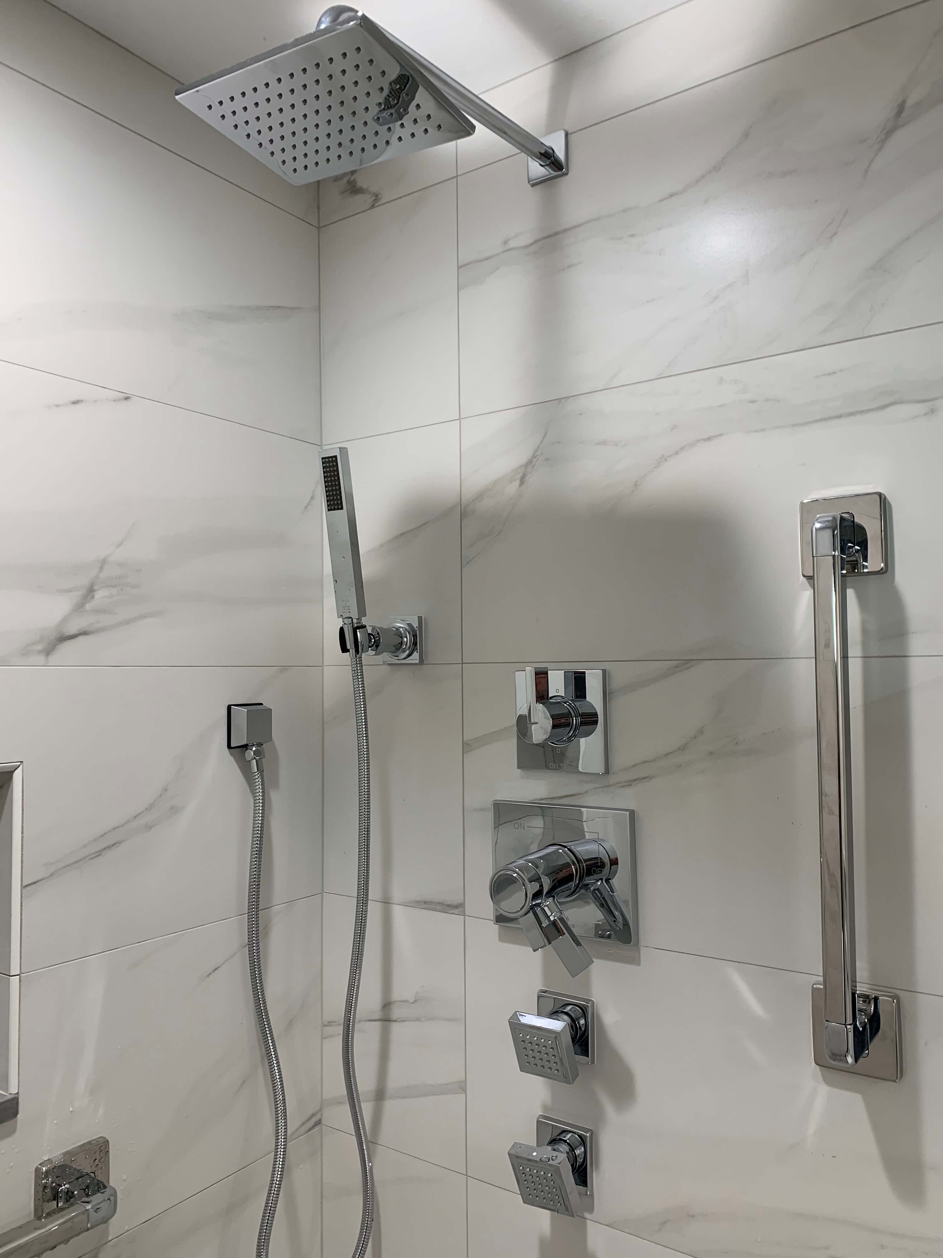 Modern shower setup with a square overhead rain showerhead, handheld shower, multiple control knobs, and a safety grab bar on white marble tiled wall.