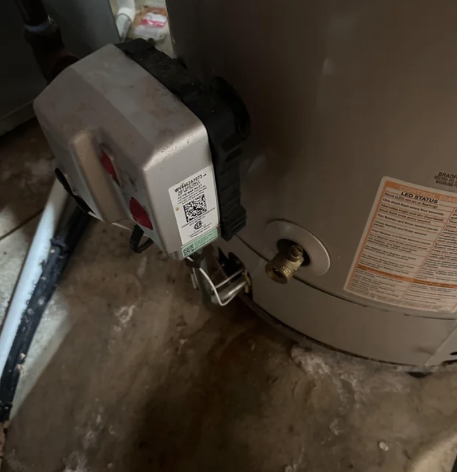 Close-up of a gas control valve and thermostat mounted on a water heater with an adjacent brass fitting and a visible LED status label.