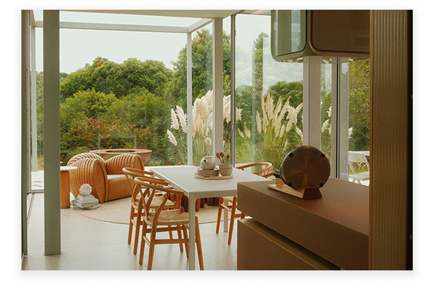 An artist's impression of a modern dining area in Gurner Grove Estate, featuring wooden chairs and a table in a glass-enclosed room with views of lush Austral, NSW greenery and pampas grass.