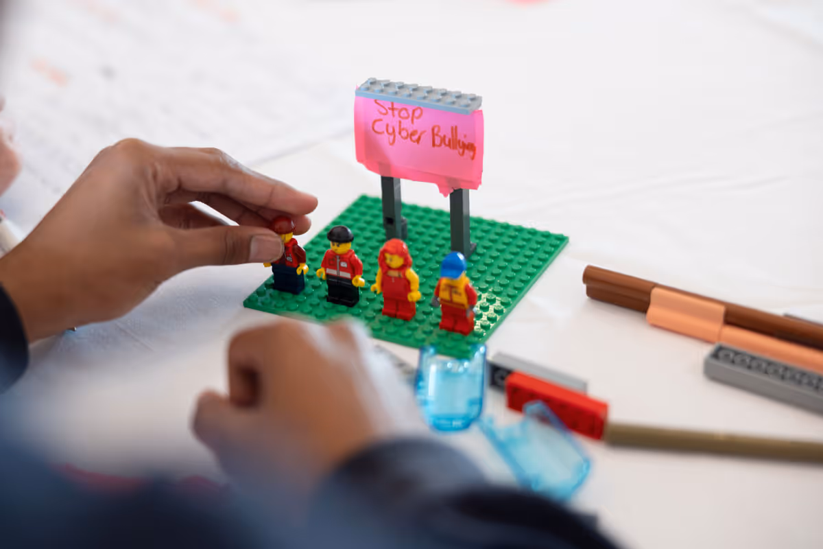 Hands arranging Lego figures on a green baseplate with a sign saying 'Stop Cyber Bullying'.