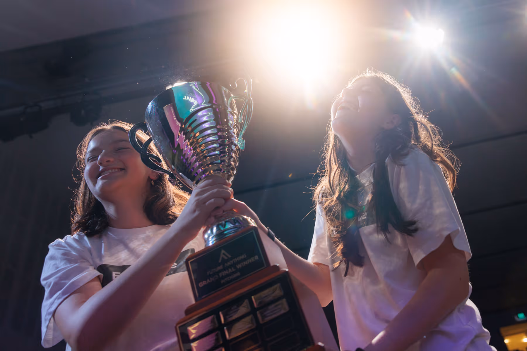 Two young women smiling and holding a large trophy under bright stage lights.
