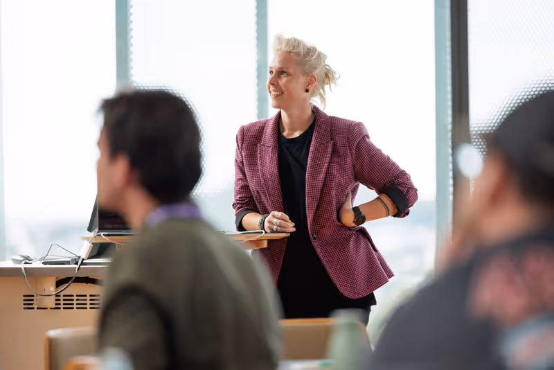 Woman in a checkered blazer standing and smiling in a bright conference room space with blurred people in the foreground.