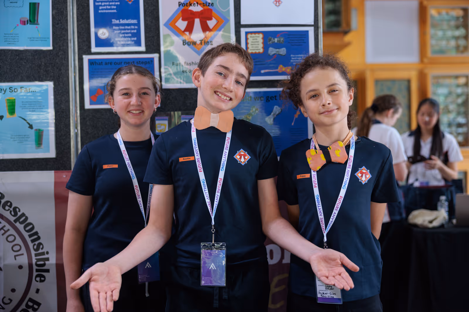 Three smiling children wearing navy shirts and lanyards standing indoors in front of a display board.