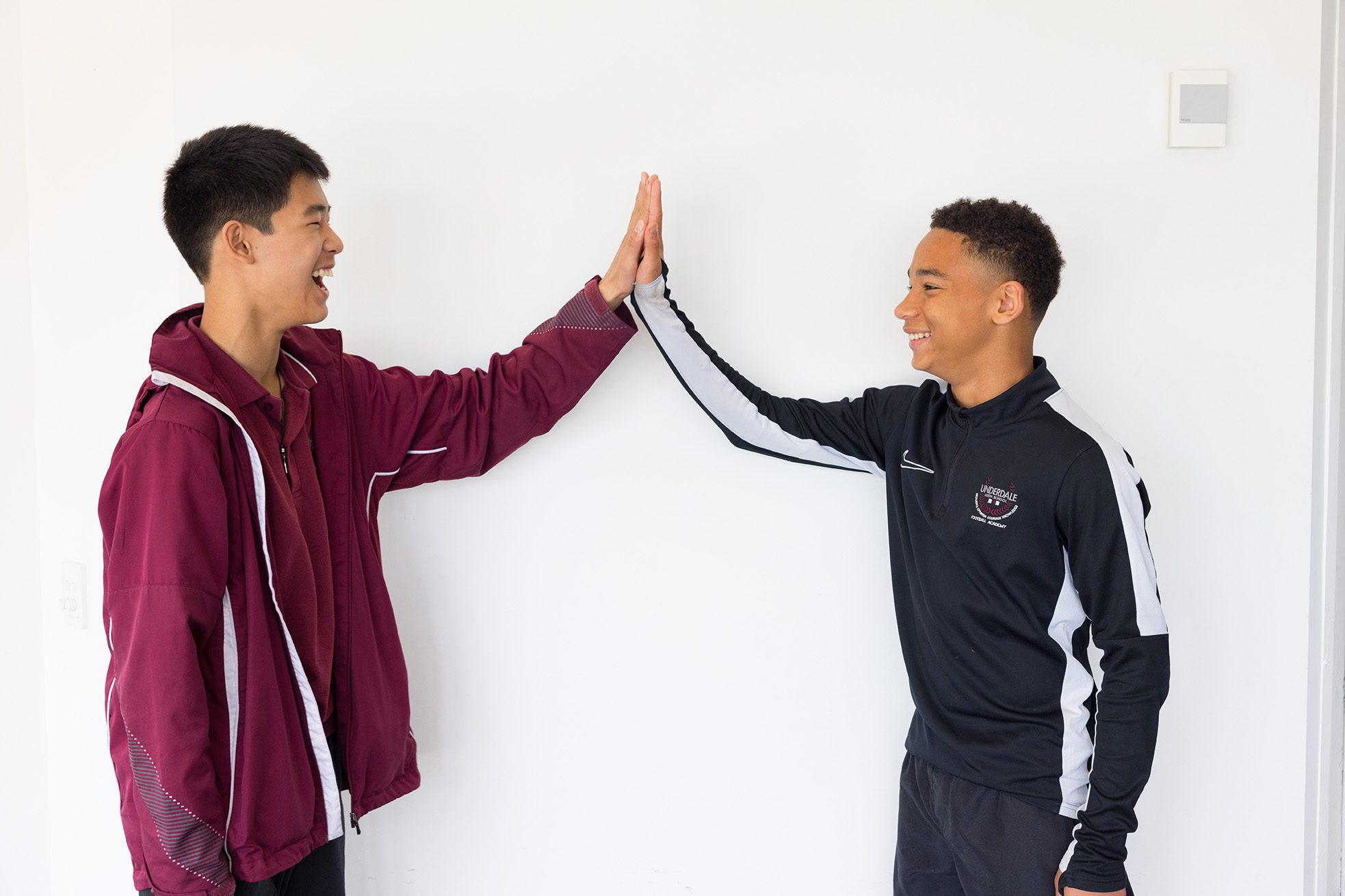 Two teenage boys smiling and giving each other a high five against a plain white wall.