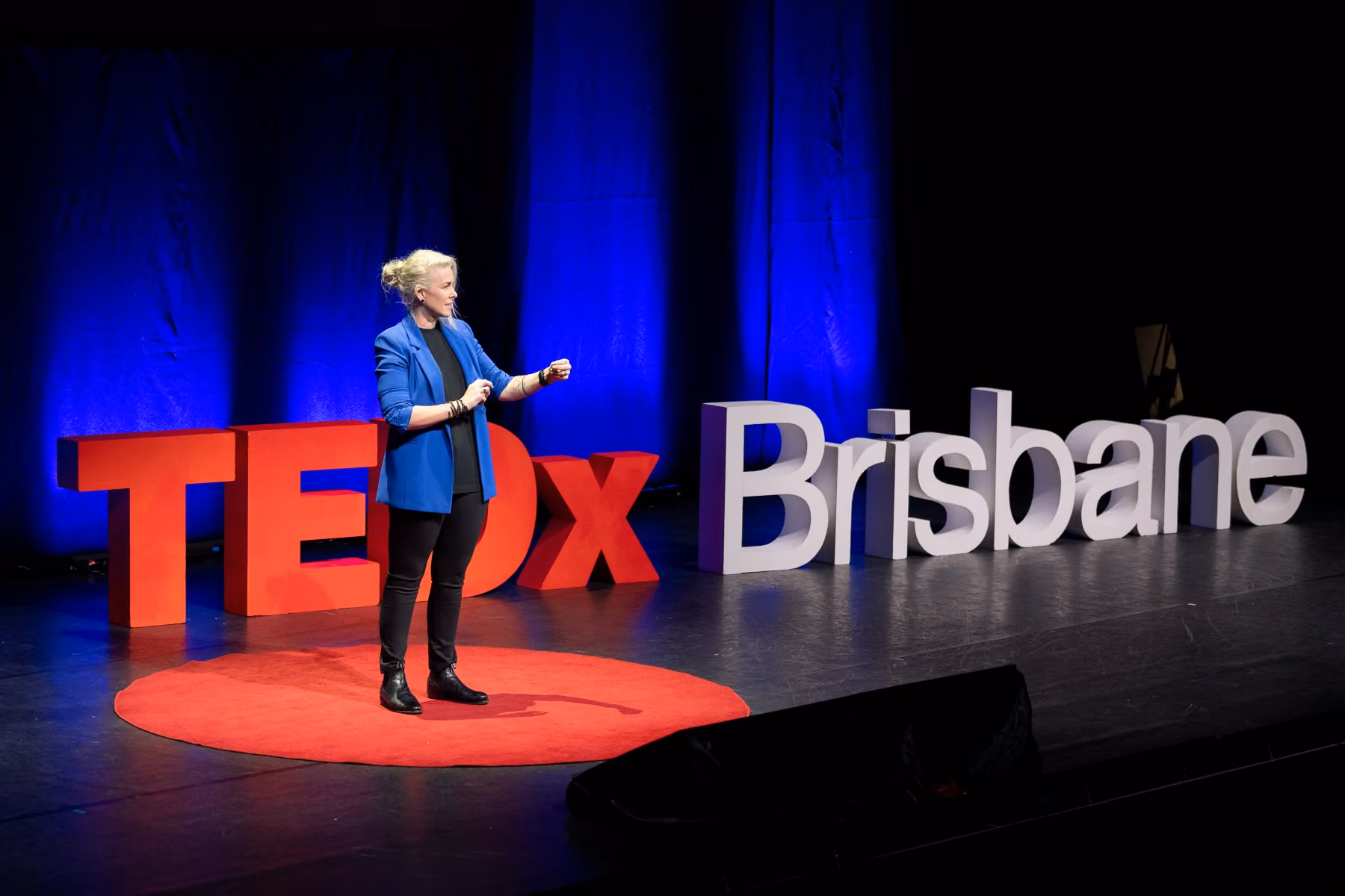 Speaker, Nicole Dyson, in blue jacket delivers a TEDx talk on stage with large TEDx Brisbane letters behind.