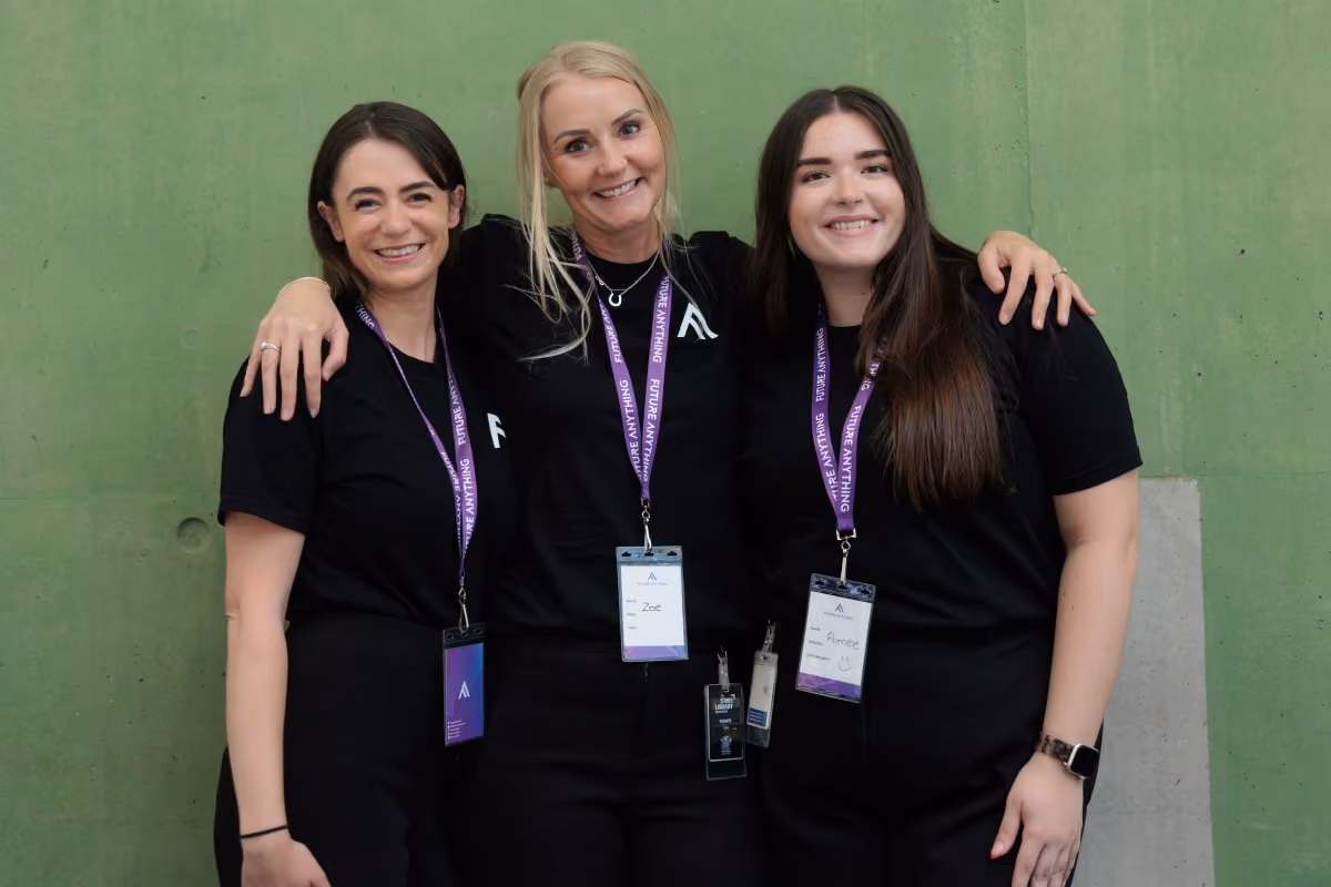 Three women standing side by side in front of a green wall, smiling and wearing black shirts with purple lanyards and name badges.