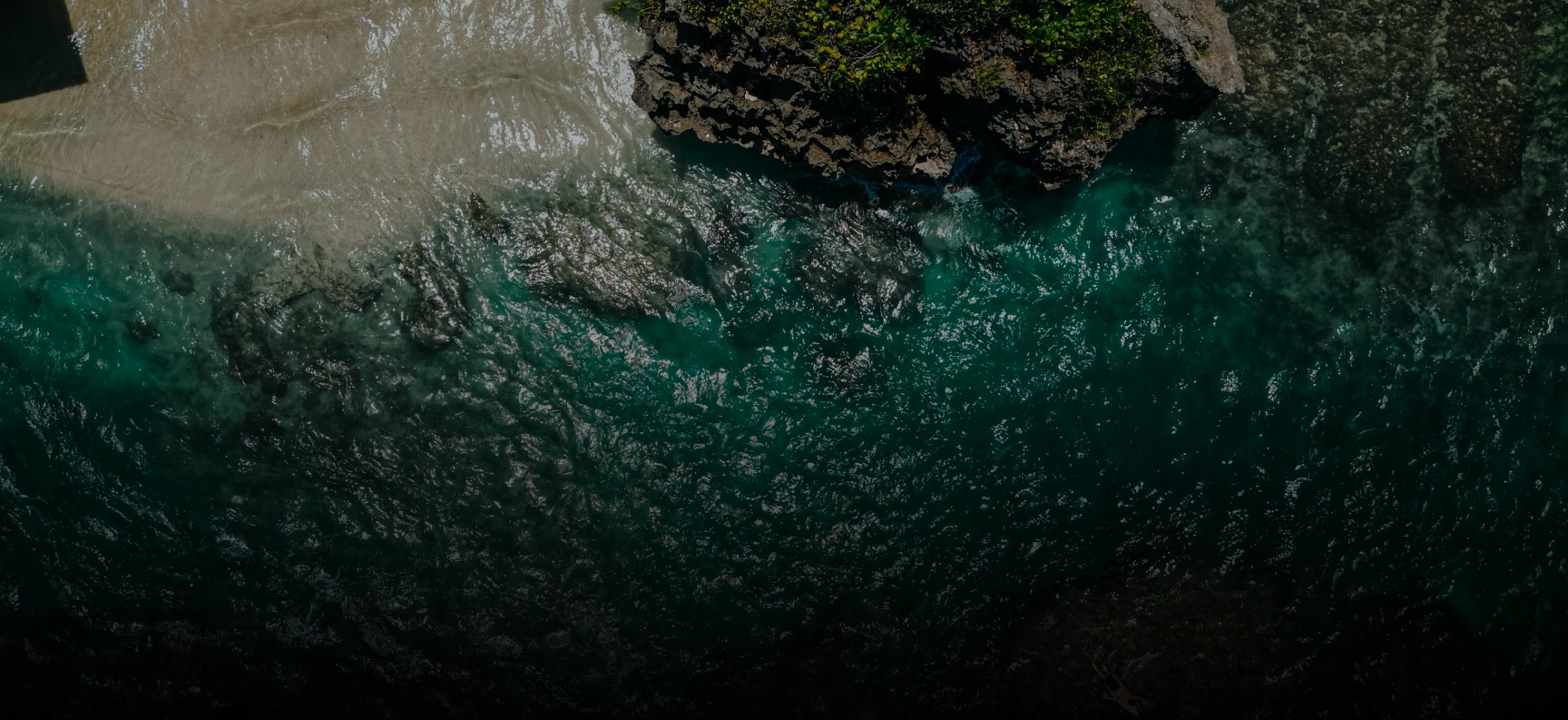 Aerial view of clear turquoise ocean water flowing around a rocky shoreline with some vegetation.