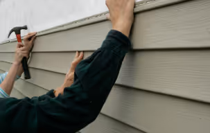 A man is using a hammer to install siding on a building, focusing intently on his task.
