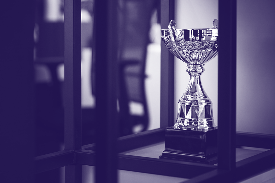 A silver trophy with intricate details is displayed on a black base inside a glass case. The background is blurred, showcasing office chairs and a modern workspace, tinted in shades of purple for a stylized effect.