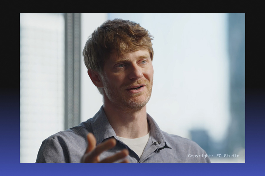 A man with light hair speaks in a bright office, gesturing with one hand while wearing a gray shirt and lapel mic, with blurred city buildings behind him.