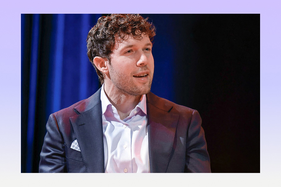 A man with curly hair in a suit jacket and open-collar shirt speaks on stage against a dark blue backdrop, appearing engaged in conversation during an event.