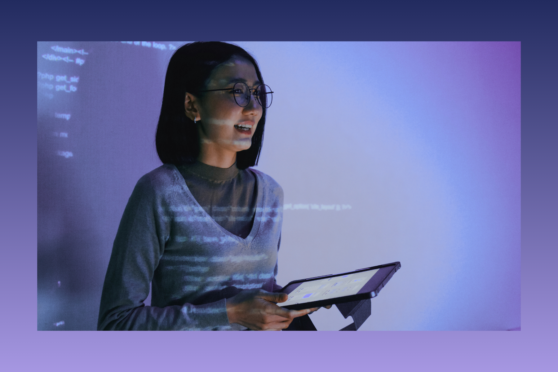 A person standing before a projected screen with code, holding a tablet and speaking, illuminated by blue and purple light.