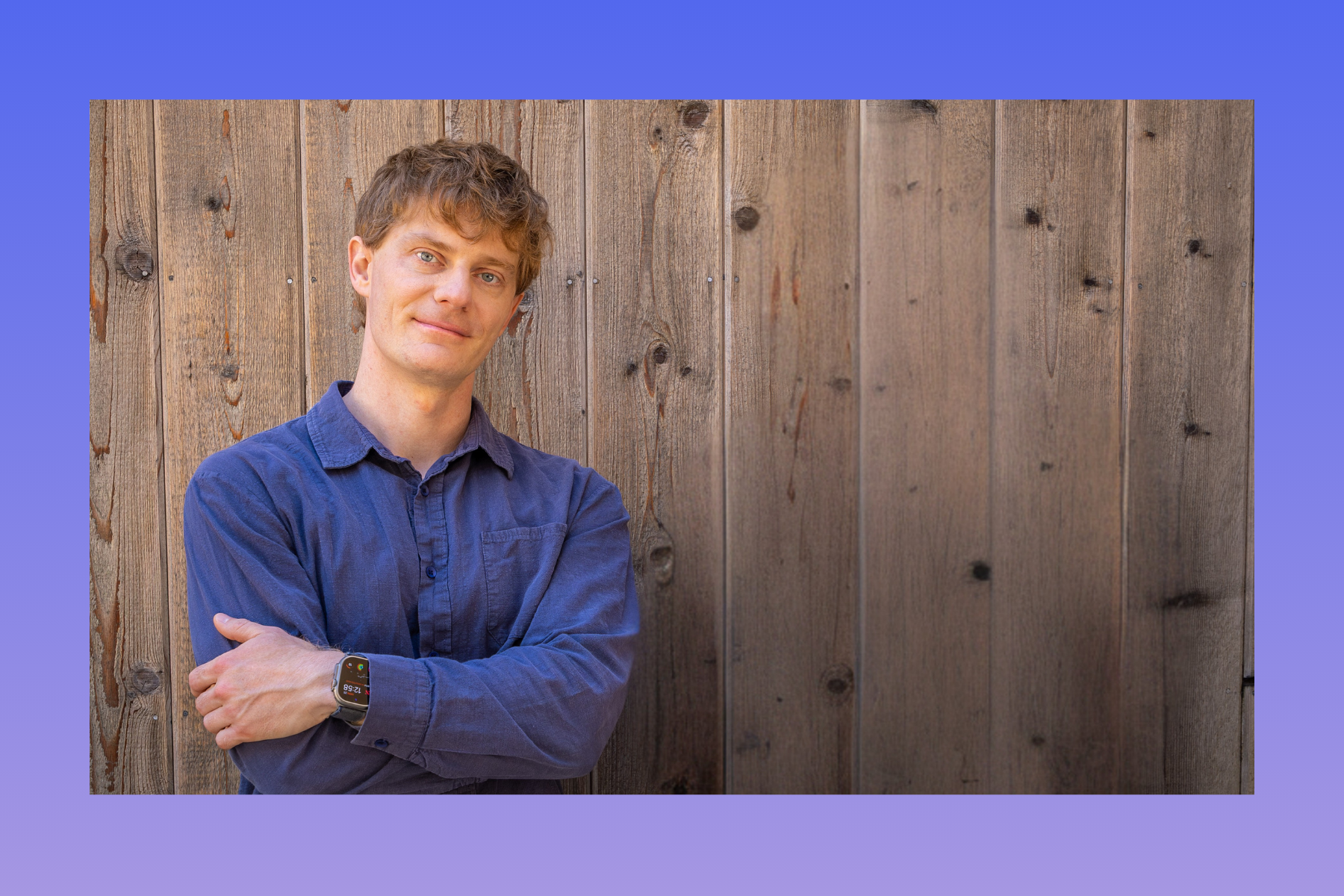 A person with short wavy hair stands smiling with arms crossed against a wooden fence, wearing a blue shirt and a smartwatch.