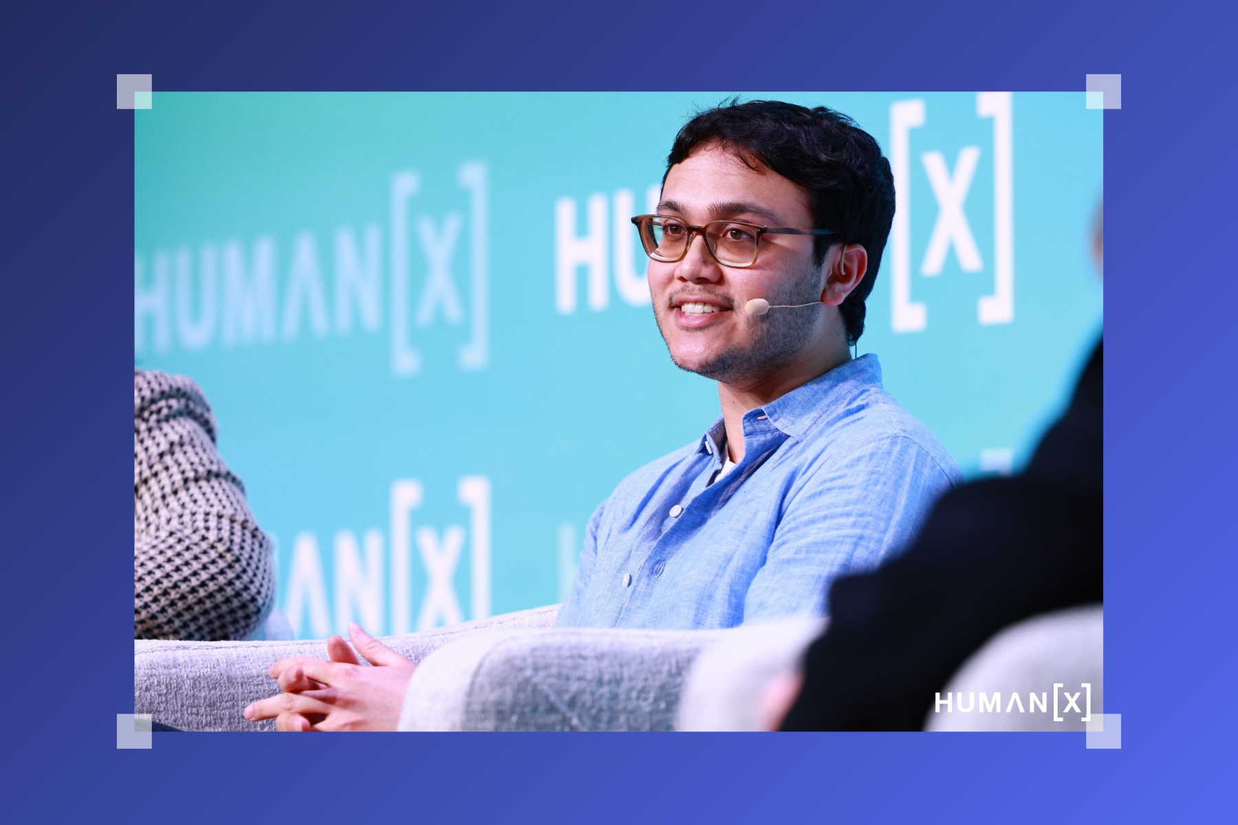 A person wearing glasses and a headset mic speaks onstage during a panel discussion, seated on a chair against a teal backdrop displaying the event’s logo.