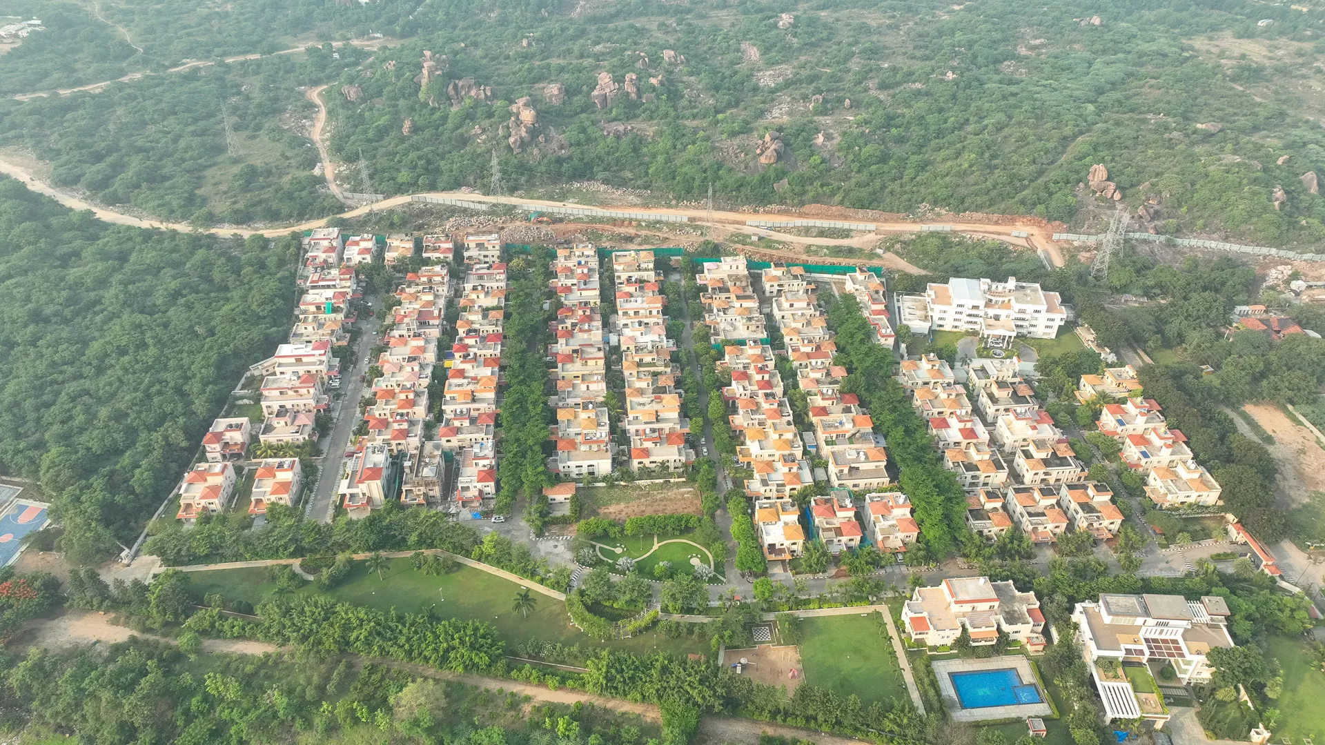 Aerial view of a residential neighborhood with rows of houses, green trees, gardens, a swimming pool, and surrounding forested hills.
