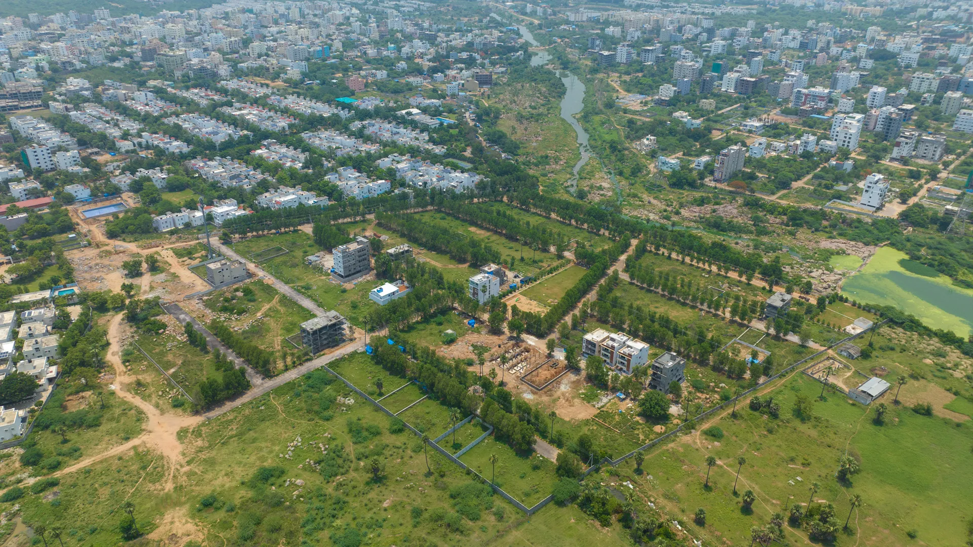 Aerial view of a suburban area with scattered residential buildings, green fields, tree-lined pathways, and a river running through the landscape.