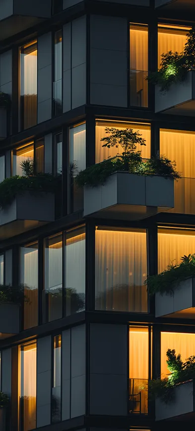 Modern building facade at dusk with illuminated windows and balconies featuring plants.