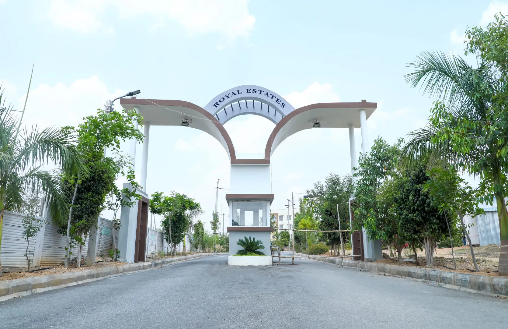 Entrance gate with curved white pillars and 'Royal Estates' sign, flanked by trees and a small guard booth in the center.