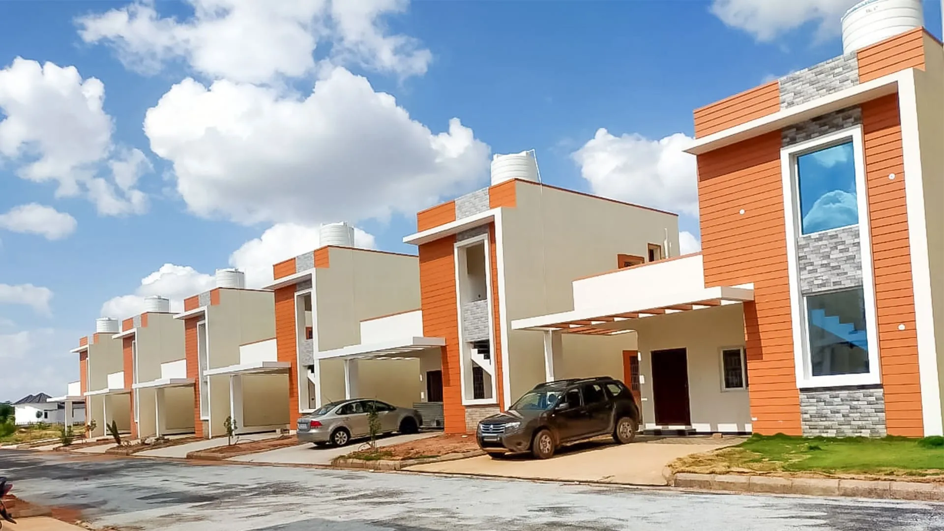 Row of modern two-story houses with orange and beige walls, covered parking, and cars parked outside on a bright sunny day.