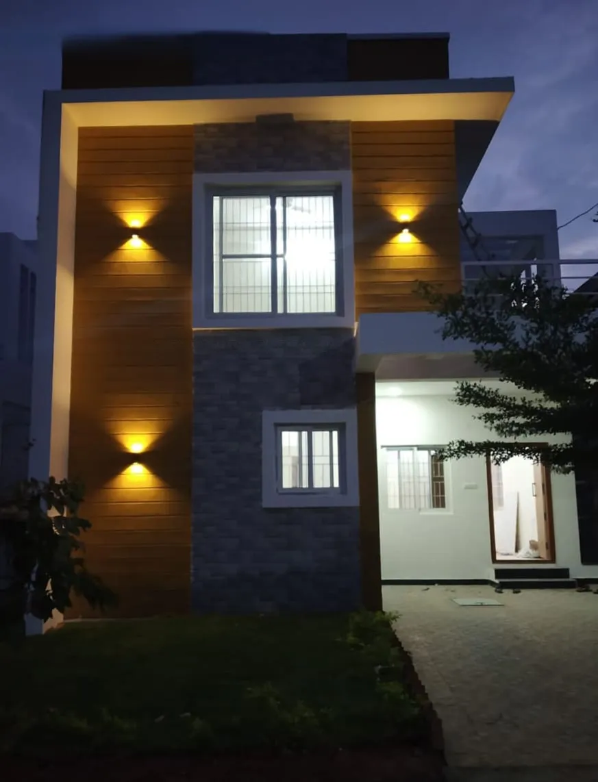 Two-story modern house exterior at dusk with warm wall lights, large windows, a driveway, and some greenery.
