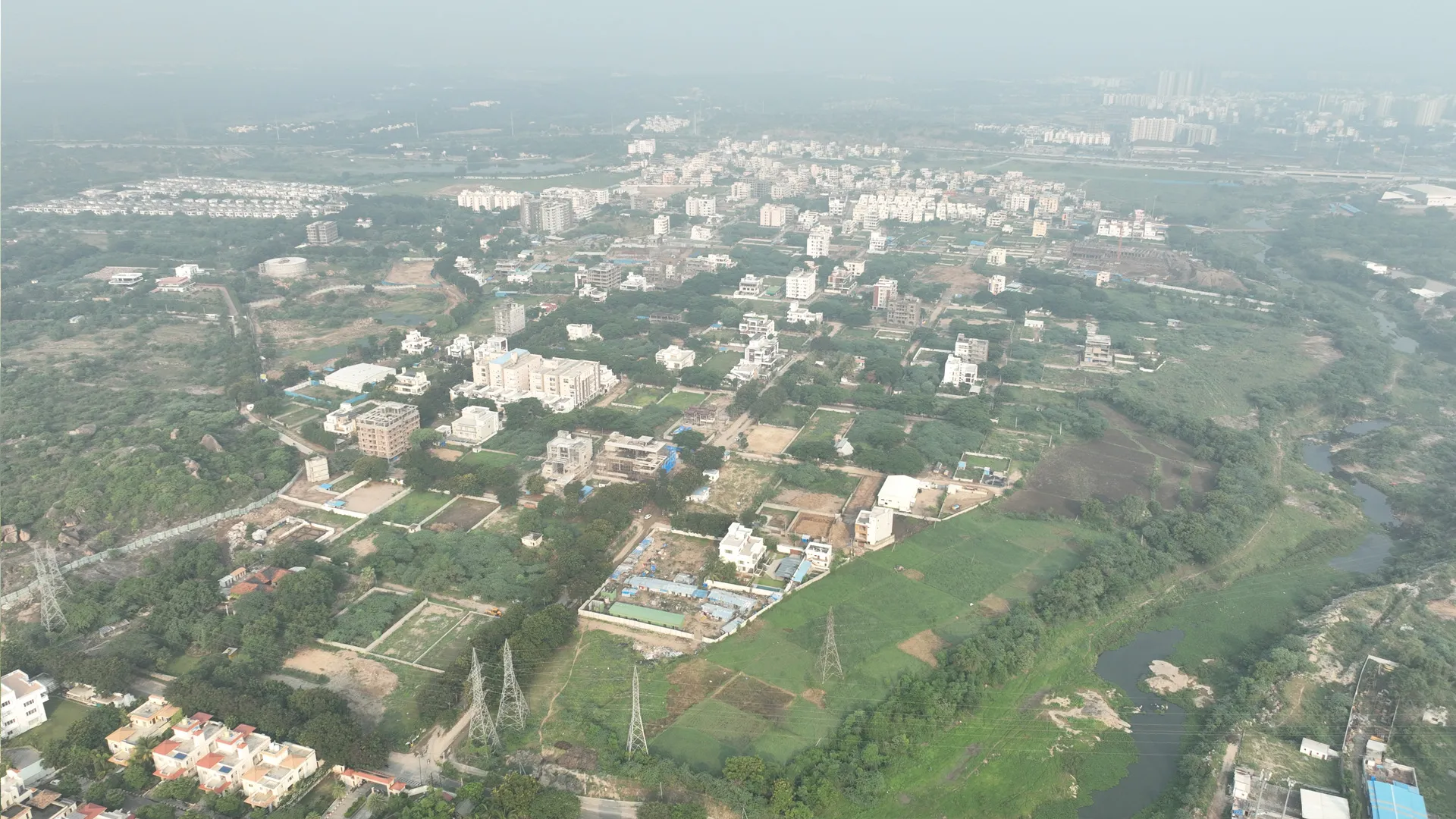 Aerial view of a suburban area with scattered buildings, green fields, and a winding river on a hazy day.