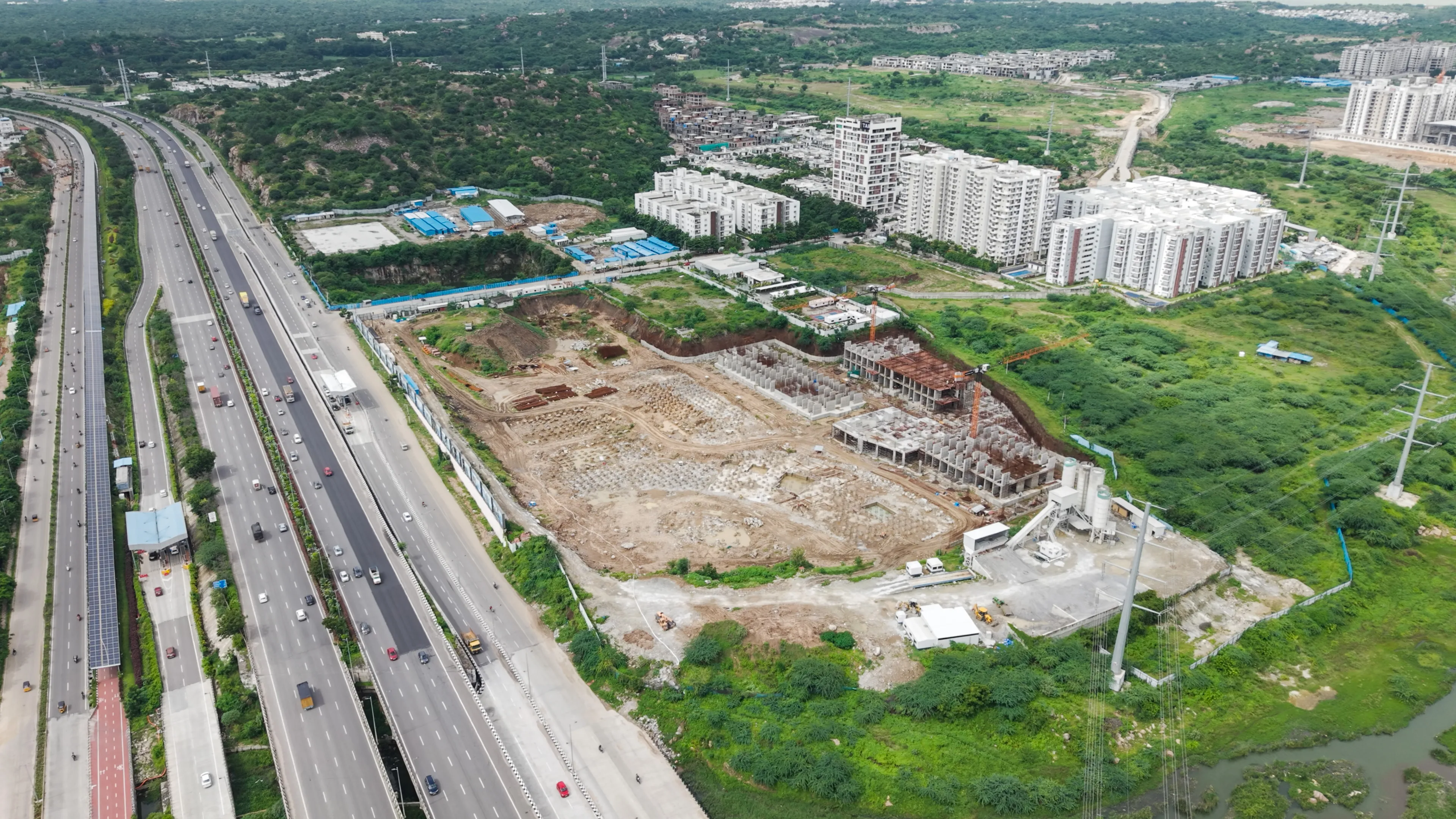 Aerial view of a highway with multiple lanes beside a large construction site and residential buildings surrounded by green fields.