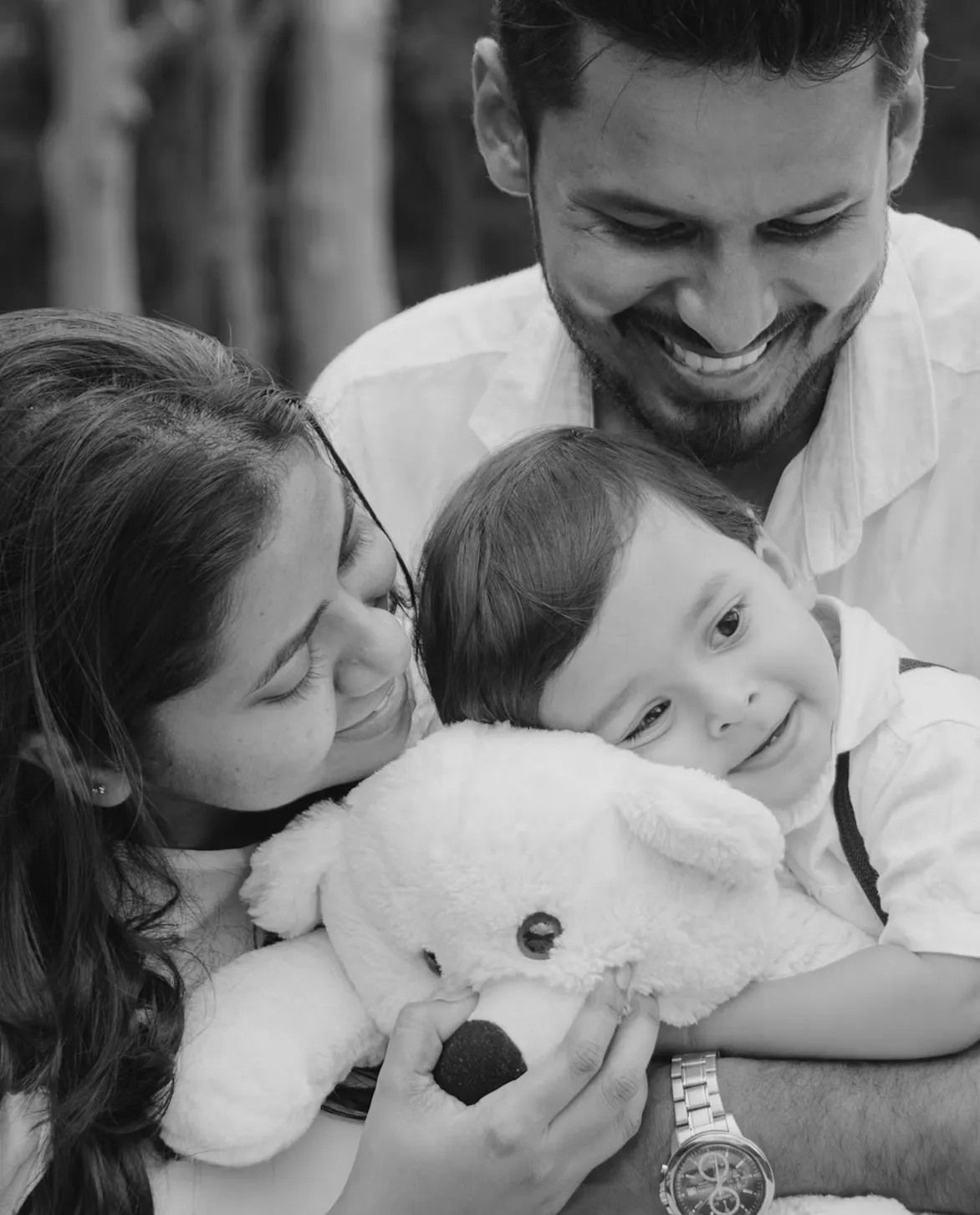 Smiling parents embracing and cuddling their young child who hugs a large stuffed teddy bear.