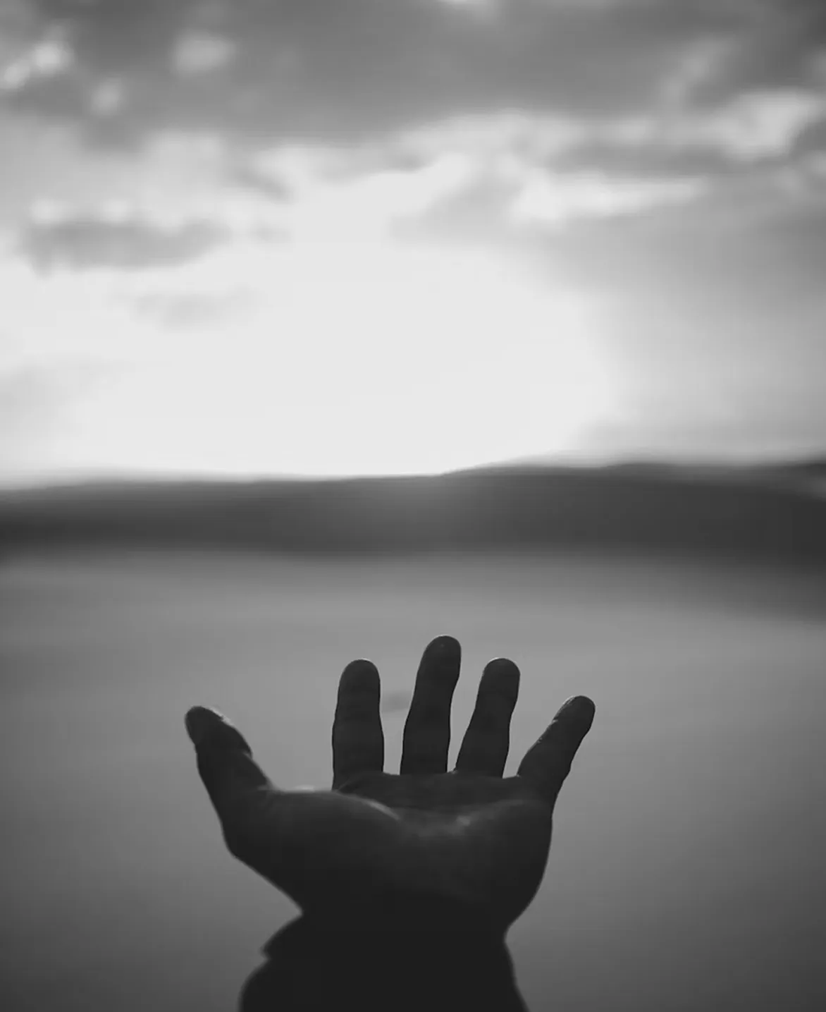 Outstretched hand reaching toward a bright sunrise or sunset over a calm landscape in black and white.
