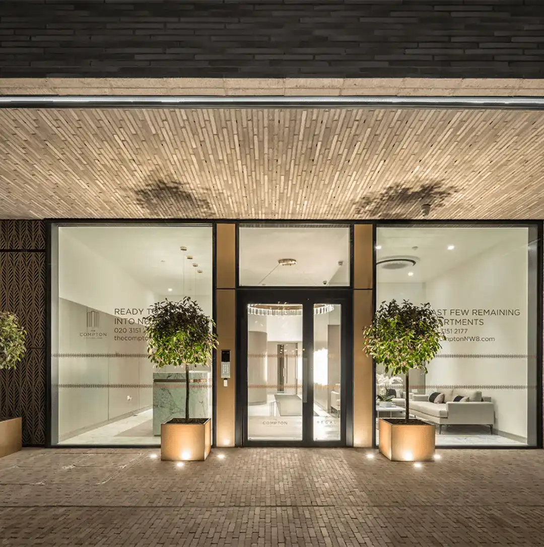 Modern brick entrance with glass doors and illuminated planters at The Compton apartments, featuring slim linear brick detailing on walls and ceiling.