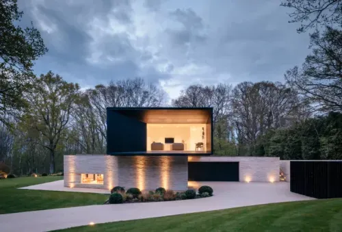 Contemporary two-storey home with light brick ground floor and dark cantilevered upper level, illuminated exterior lighting and landscaped driveway at dusk.