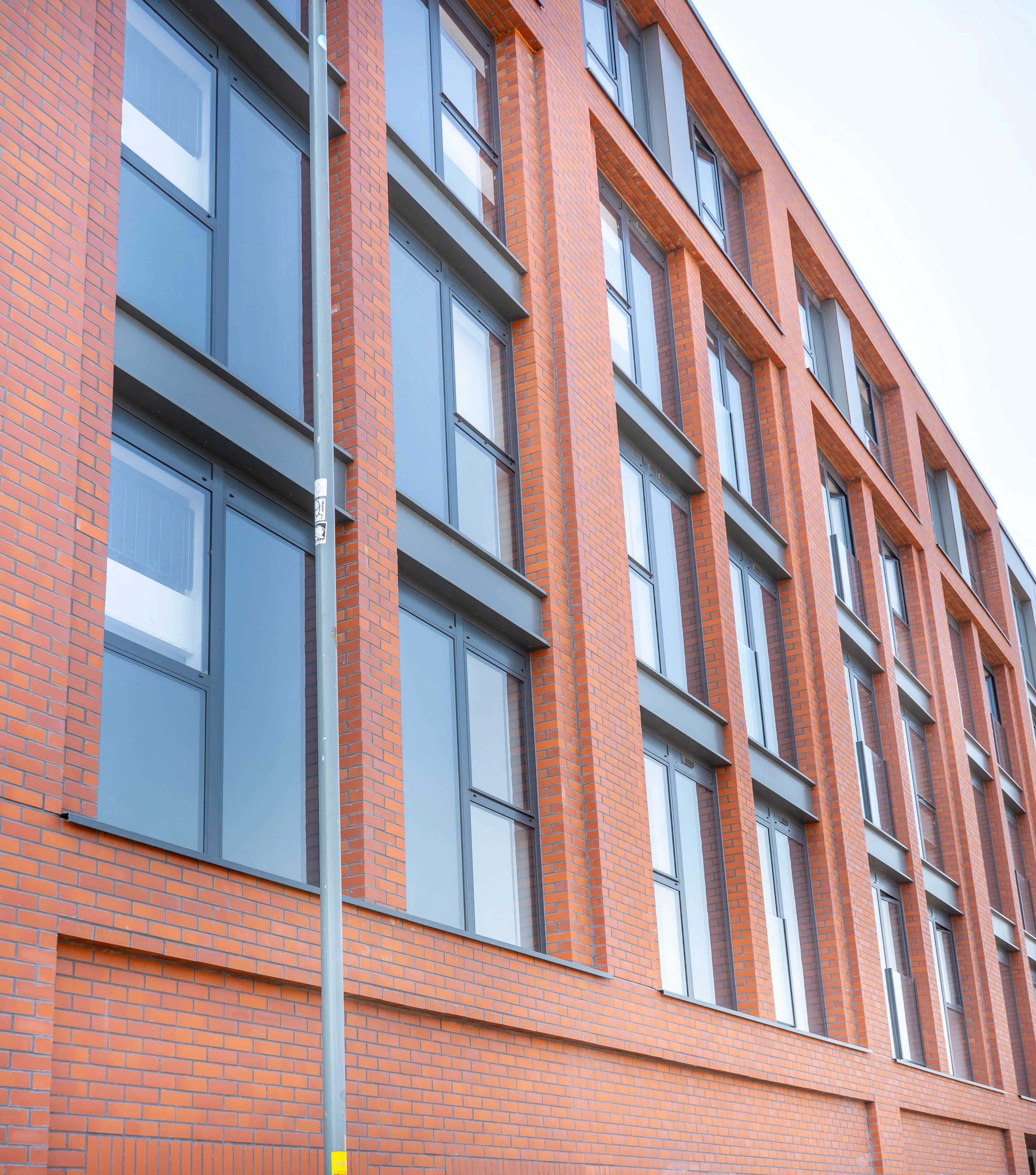 Modern red brick building with large vertical windows and grey aluminium frames, viewed from street level against a clear sky.