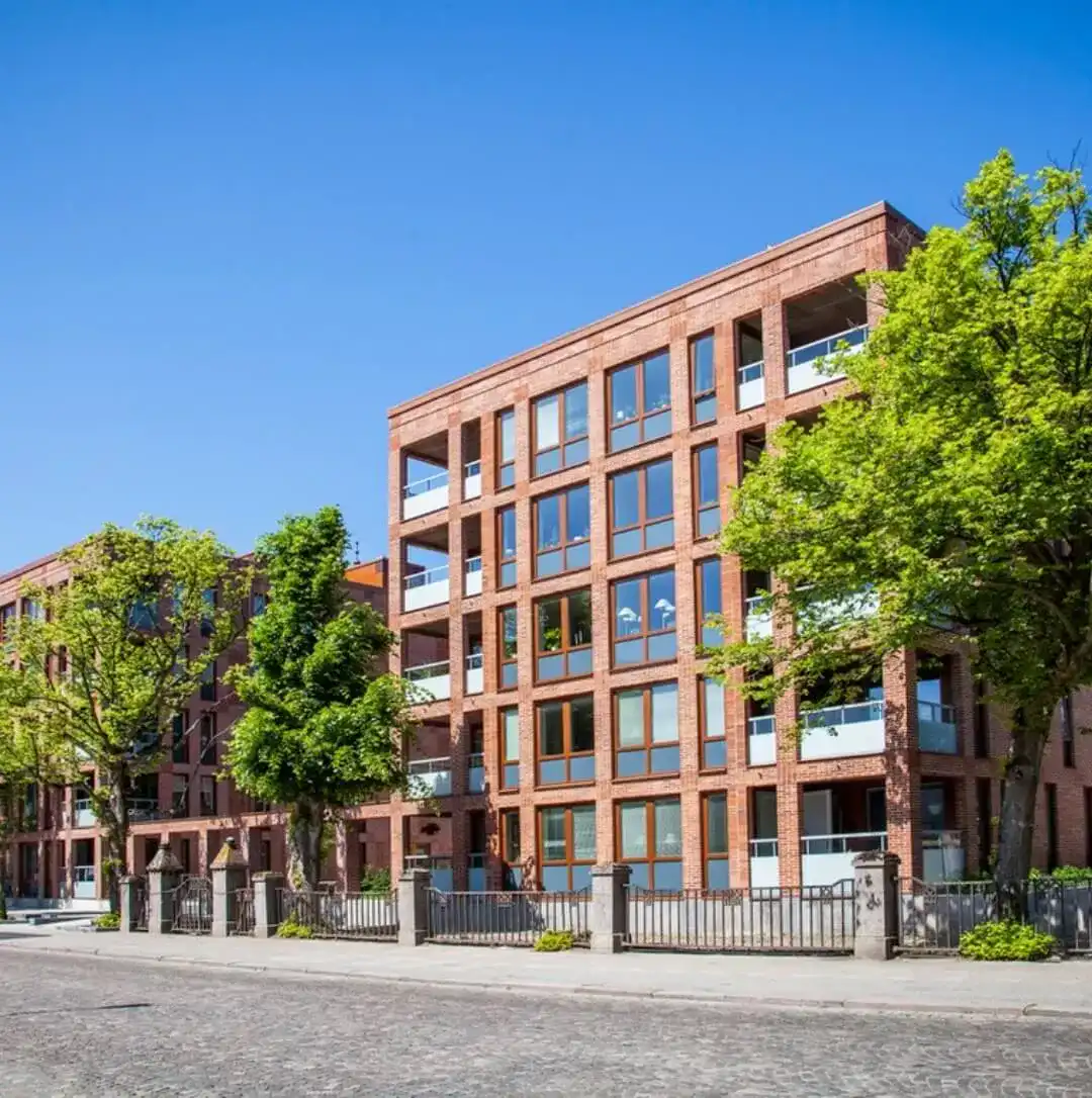 Modern red brick apartment buildings with balconies and large windows, lined with trees along a quiet city street under a clear blue sky.