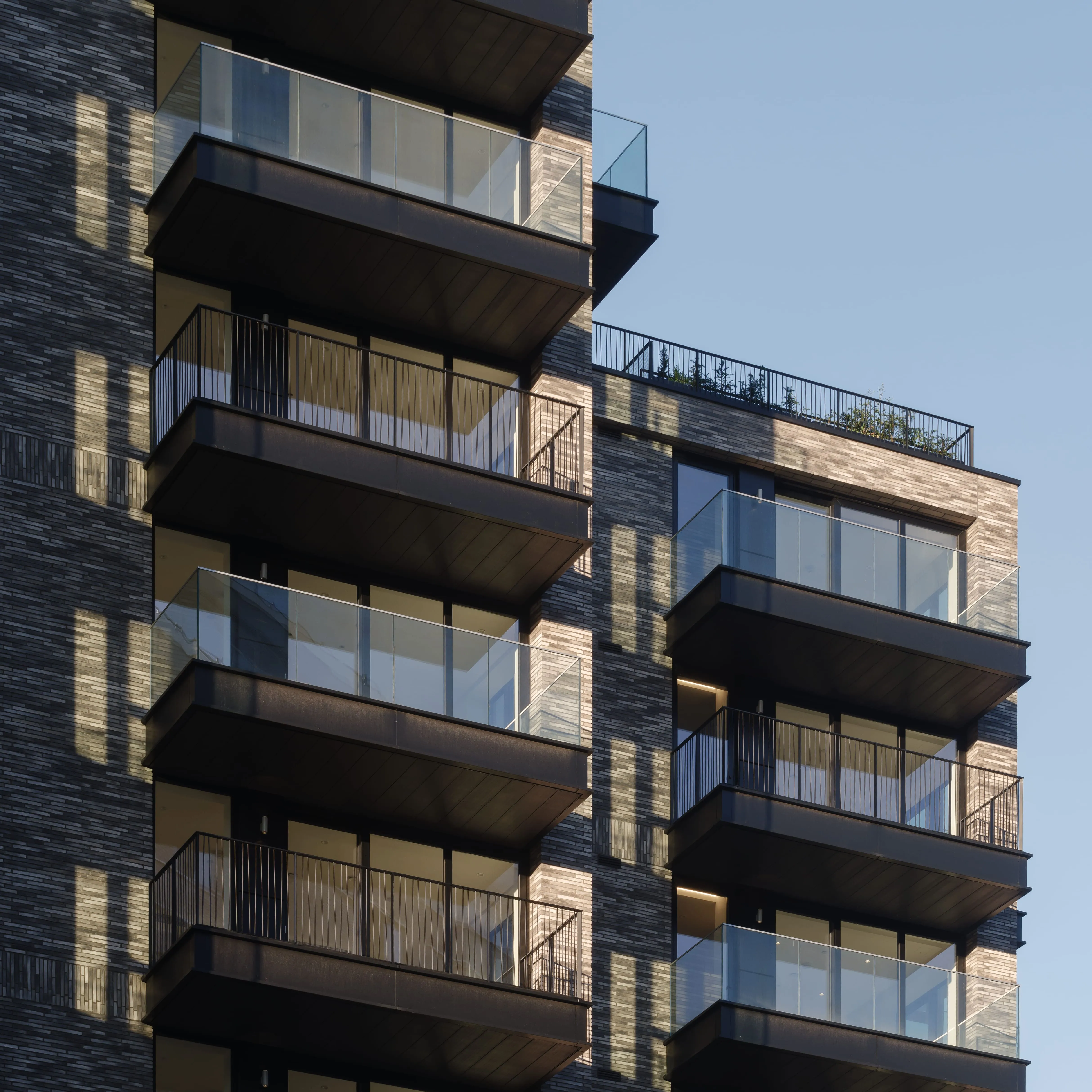 Close-up of dark brick apartment façade featuring glass-fronted balconies with metal railings in evening light.