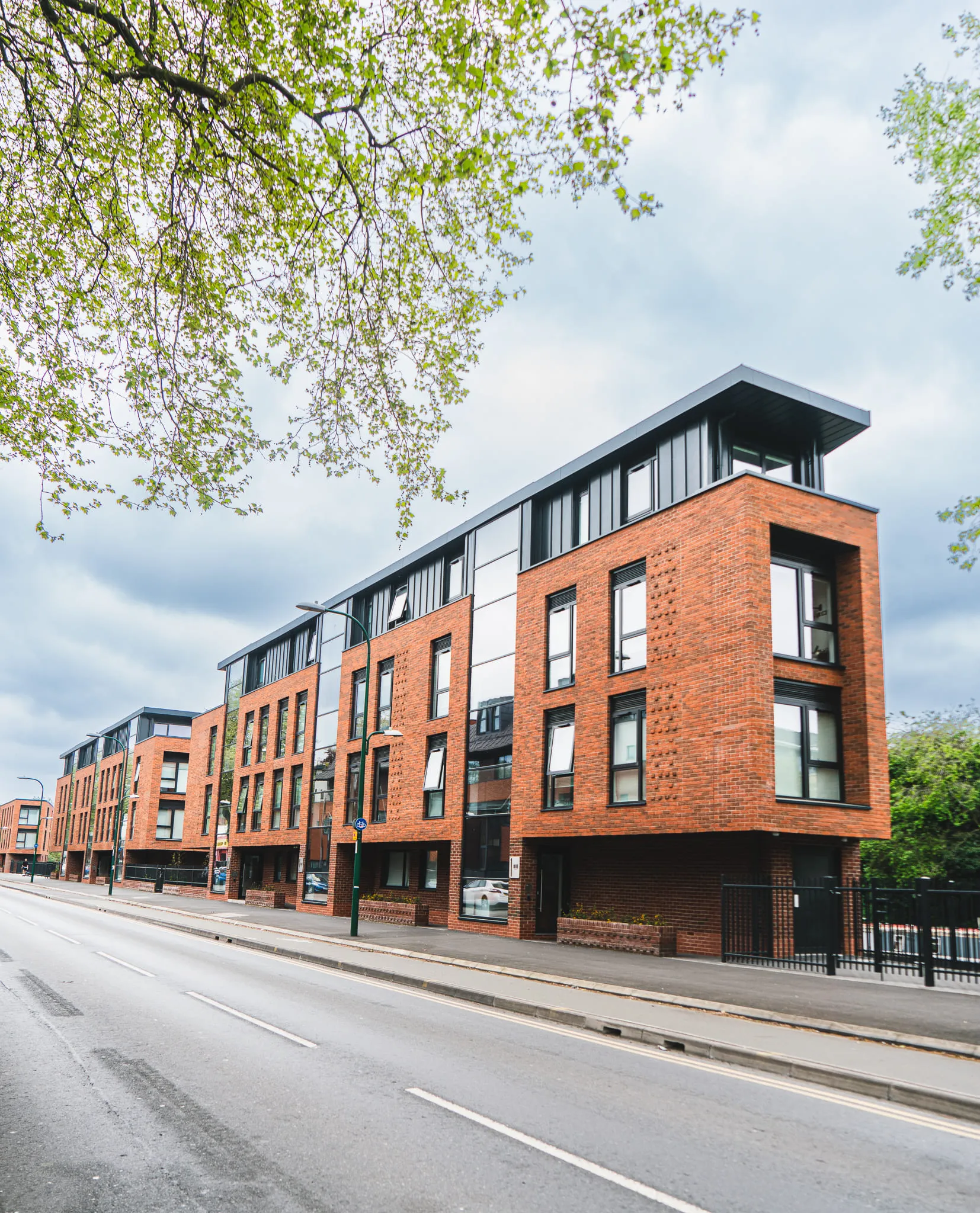 Row of modern red brick apartment buildings with large windows and dark roof extensions along a city street lined with trees.
