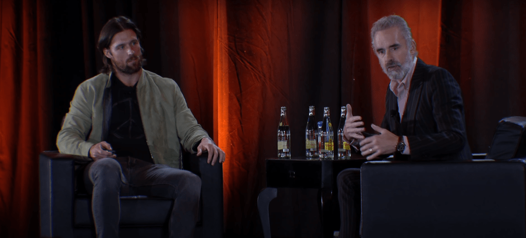 Two men seated in black chairs on stage engaged in conversation, with a small table holding five bottles between them and a dark red curtain backdrop.