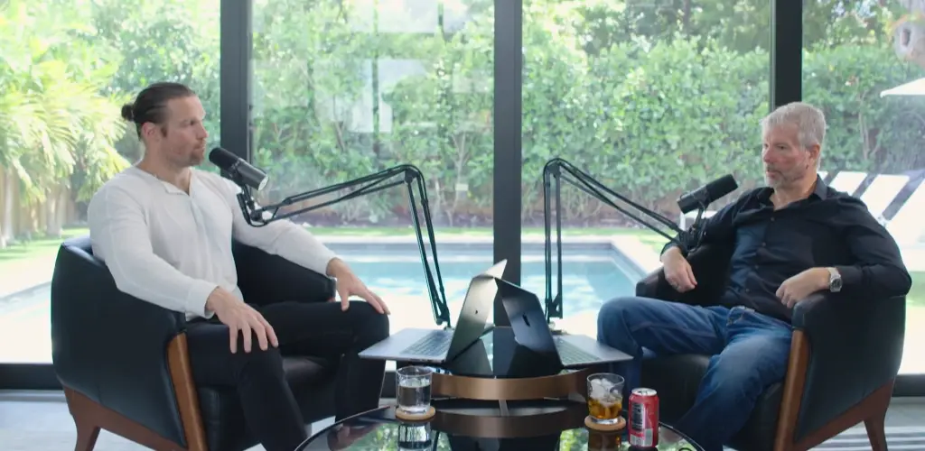 Two men seated in black armchairs facing each other, recording a podcast with microphones and laptops in a room overlooking a pool and greenery.