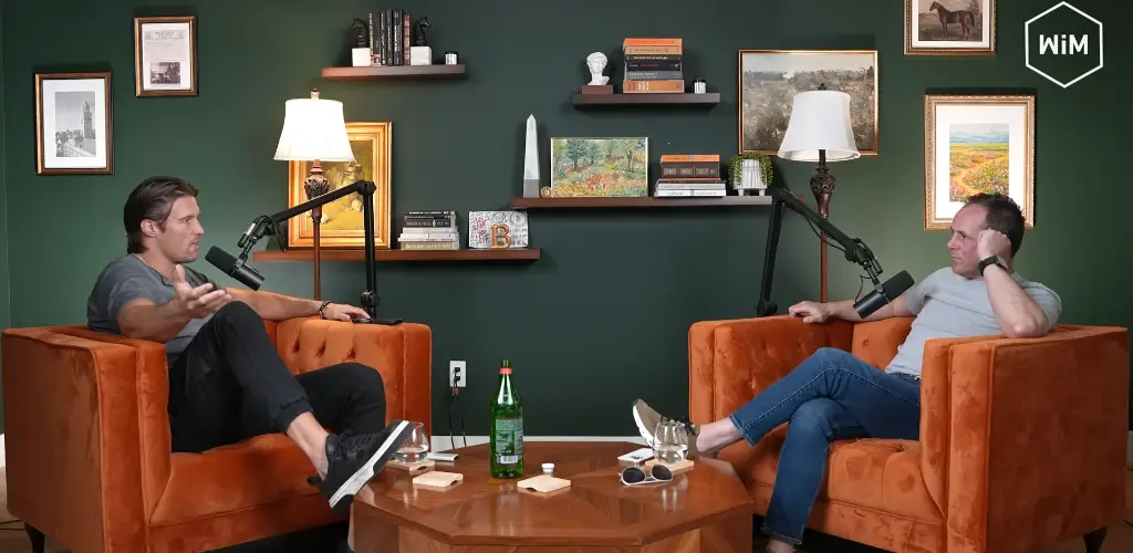 Two men seated in orange armchairs facing each other, engaged in a podcast recording with microphones in a cozy room with dark green walls and framed artwork.