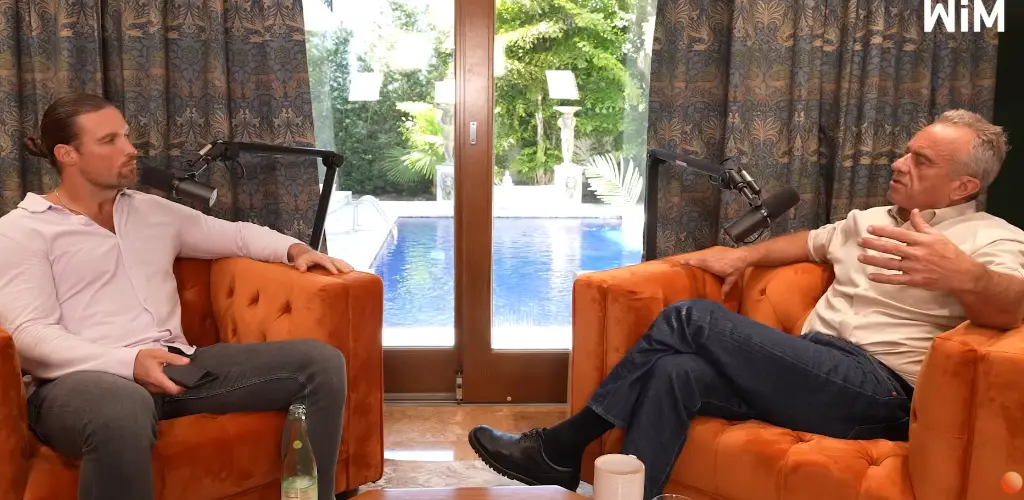 Two men having a conversation while seated on orange armchairs with a pool visible through glass doors behind them.