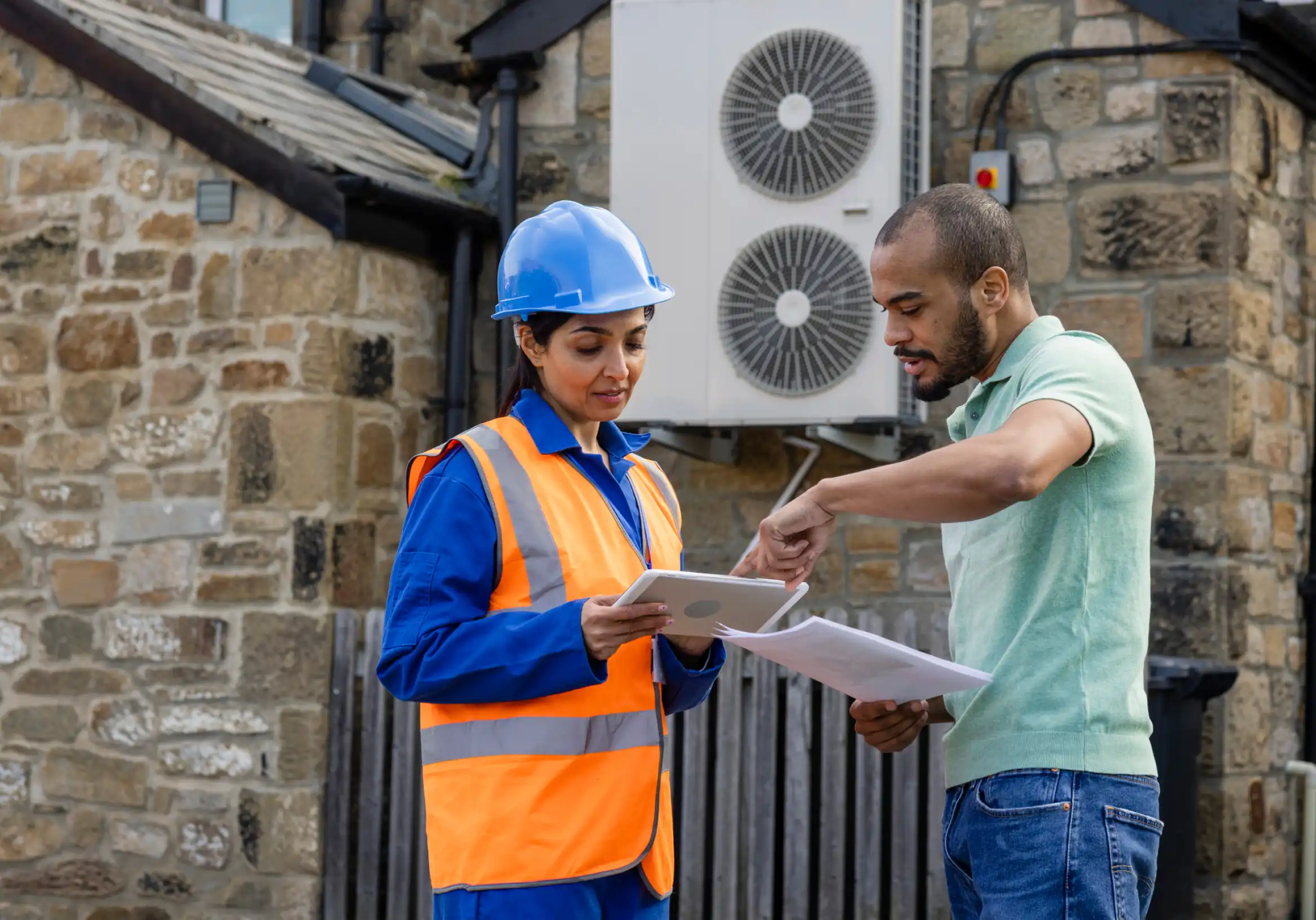 A man and a woman in hardhats looking discussing heat pump installation with a heat pump in the background.