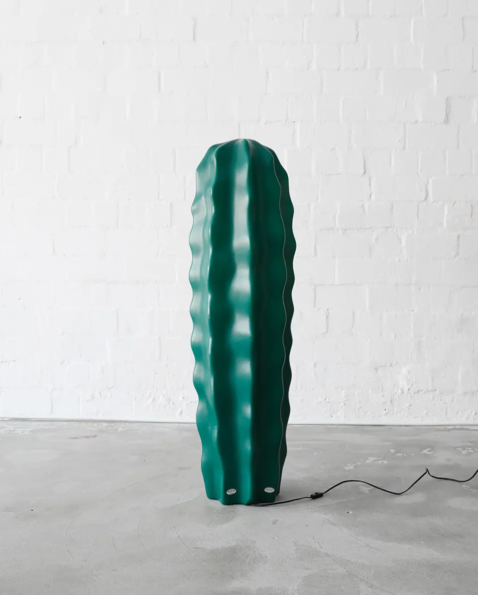 Tall cactus-shaped floor lamp in dark green color, unplugged, placed against a white brick wall