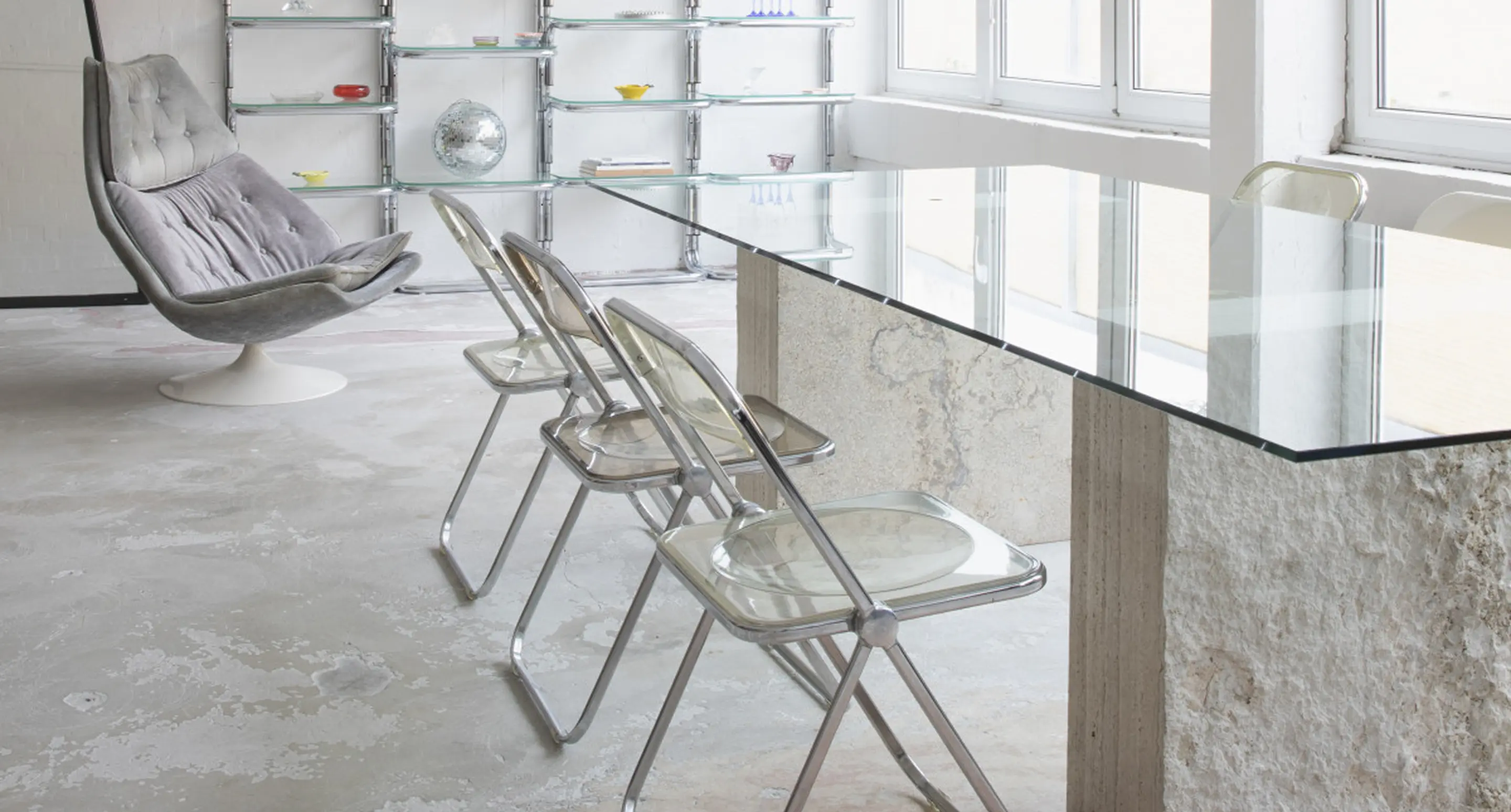 Clear folding chairs with chrome frames around a minimalist glass dining table in a bright loft