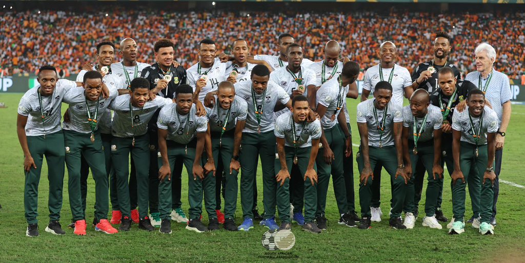 Bafana Bafana players and coaching staff pose for a celebratory team photo on the pitch, wearing white jerseys and green pants. Most team members display medals around their necks, signaling recent tournament success. The group stands united in front of a 