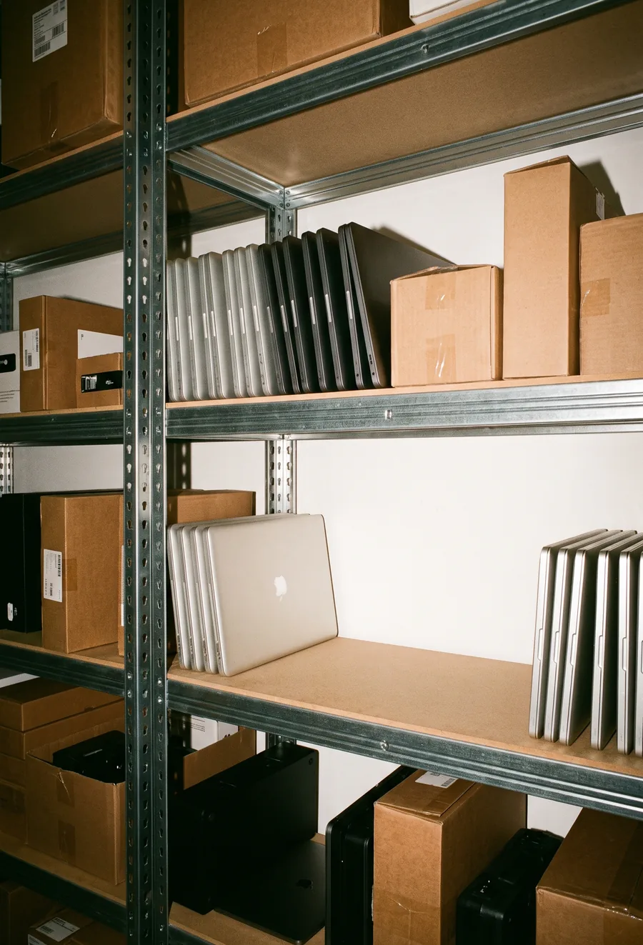 Estantería de oficina con laptops cerradas alineadas y cajas, con un hueco vacío en una balda