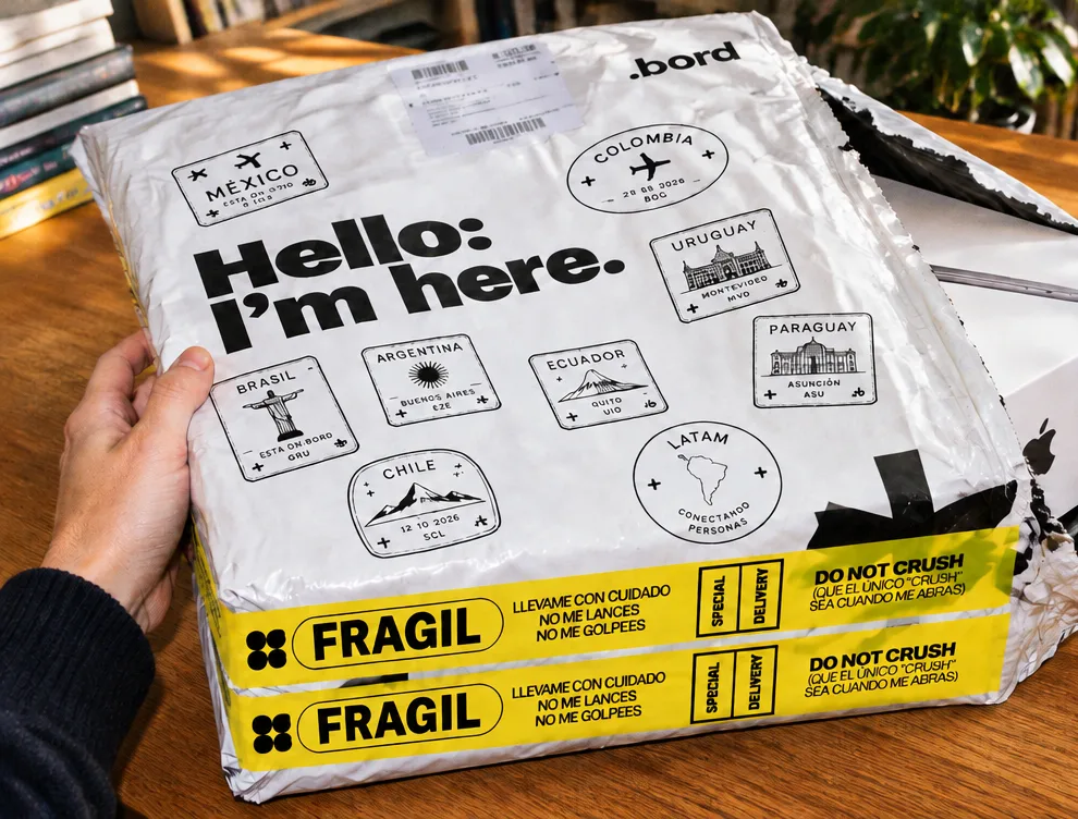 Una mano sostiene sobre un escritorio la bolsa de envío blanca de Bord, con sellos de países y cinta amarilla de frágil, de la que asoma la caja de una MacBook