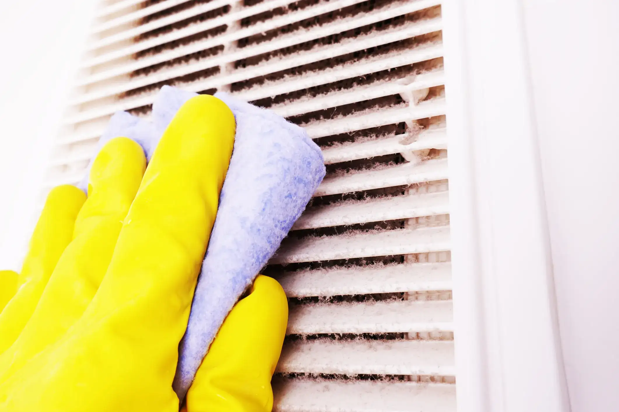 Yellow-gloved hand wiping a dusty white air vent with a blue cleaning cloth.