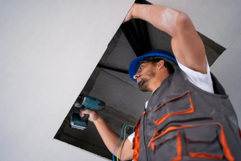 A construction worker wearing a hard hat and safety vest uses a power drill while working inside a ceiling access hole.