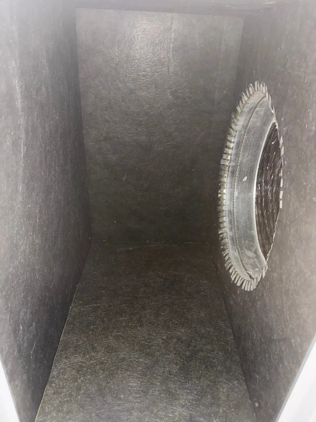 Interior Of A Rectangular Duct Or Air Plenum, Likely Part Of An HVAC System. The Walls And Floor Are Lined With A Dark Gray Or Black Fibrous Insulation Material That Has A Slightly Coarse Texture. On The Right Wall, There's A Circular Metal Takeoff Collar Or Boot, Which Is The Connection Point For A Round Duct. This Collar Has A Serrated Or Toothed Edge On Its Perimeter And A Dark, Gridded Mesh Or Damper Visible Within Its Opening. The Perspective Suggests The Viewer Is Looking Down The Length Of The Square-Cornered, Insulated Channel.