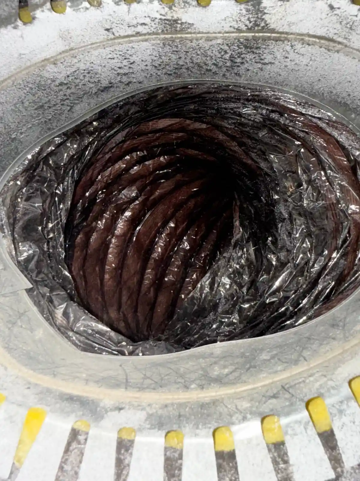 Internal View Of A Flexible HVAC Duct Connected To A Galvanized Metal Collar. The Collar Is Circular, Weathered, And Has A Ring Of Yellow Tabs Or Fins Around The Outside Edge In The Foreground. Inside The Collar, The Flexible Duct Is Visible. It Has A Crinkled, Black Or Dark Gray Plastic Outer Layer (Vapor Barrier/Jacket). Beneath This Outer Layer, The Inner Liner Appears To Be A Dark Reddish-Brown Or Black Material, Which Is Heavily Corrugated And Creates A Spiraling, Tunnel-Like Effect Leading Into The Darkness. This Internal Brown Layer Is Likely The Main Air Passage Of The Duct.