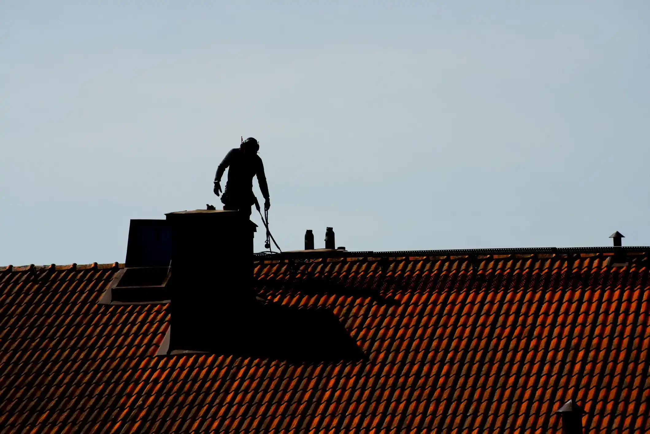 Silhouette of a chimney sweep working on a rooftop with a red tile roof and chimney stack.