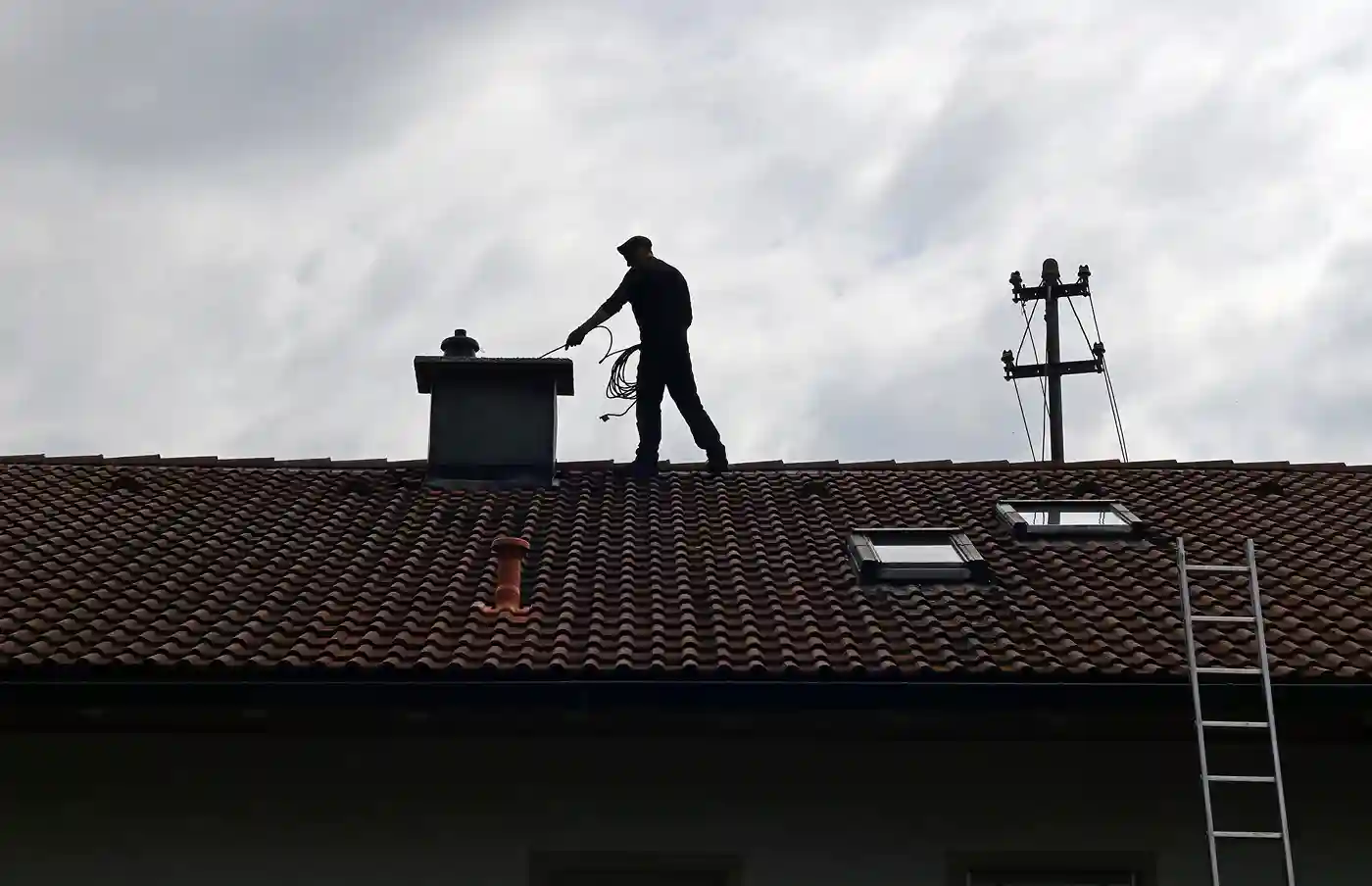 Silhouette of a chimney sweep standing on a roof, lowering a cleaning tool into the chimney stack.